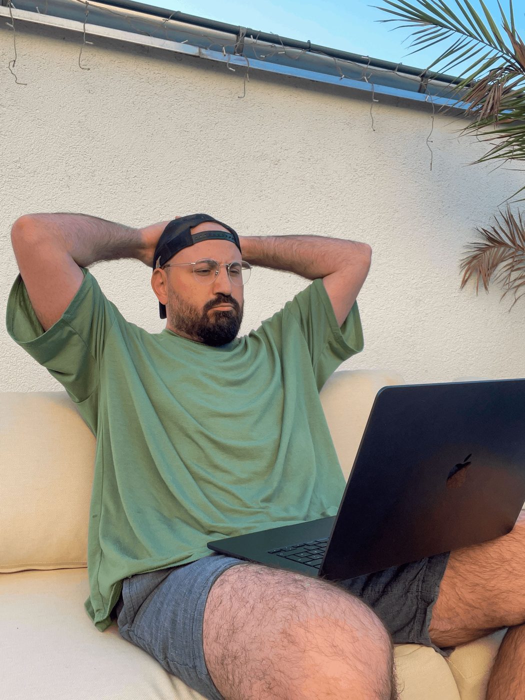 Buri working on a laptop outdoors on a sofa under a palm tree
